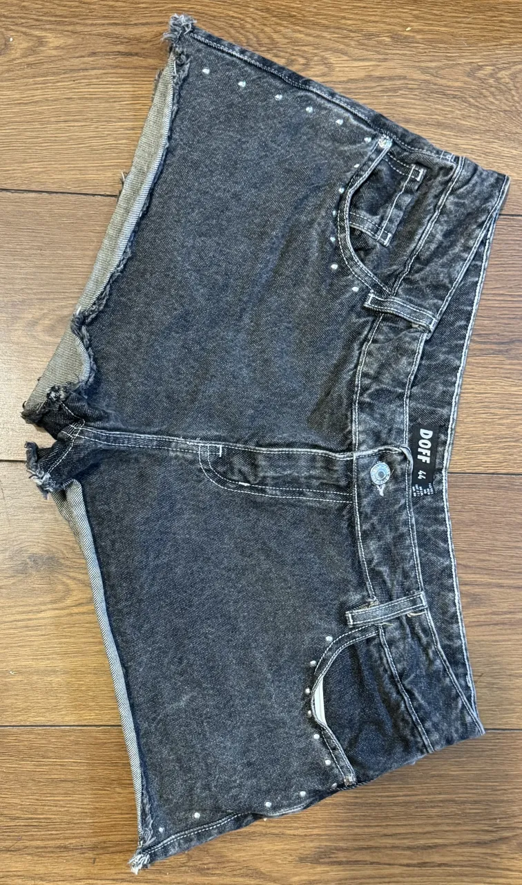Shorts de jean negros con detalles de tachas plateadas en los bolsillos delanteros y traseros. Los bordes de las piernas son desflecados.