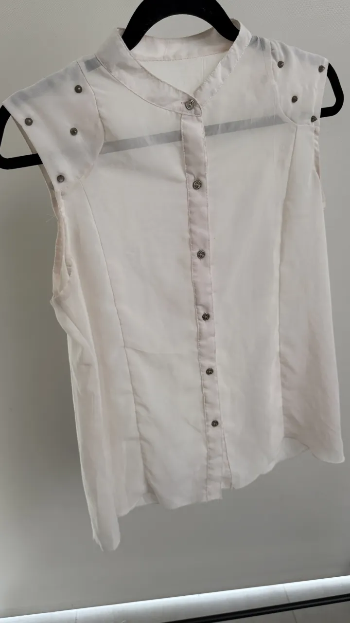 Remera blanca sin mangas con cuello mao y botones al frente. Detalle de tachas metálicas en los hombros.