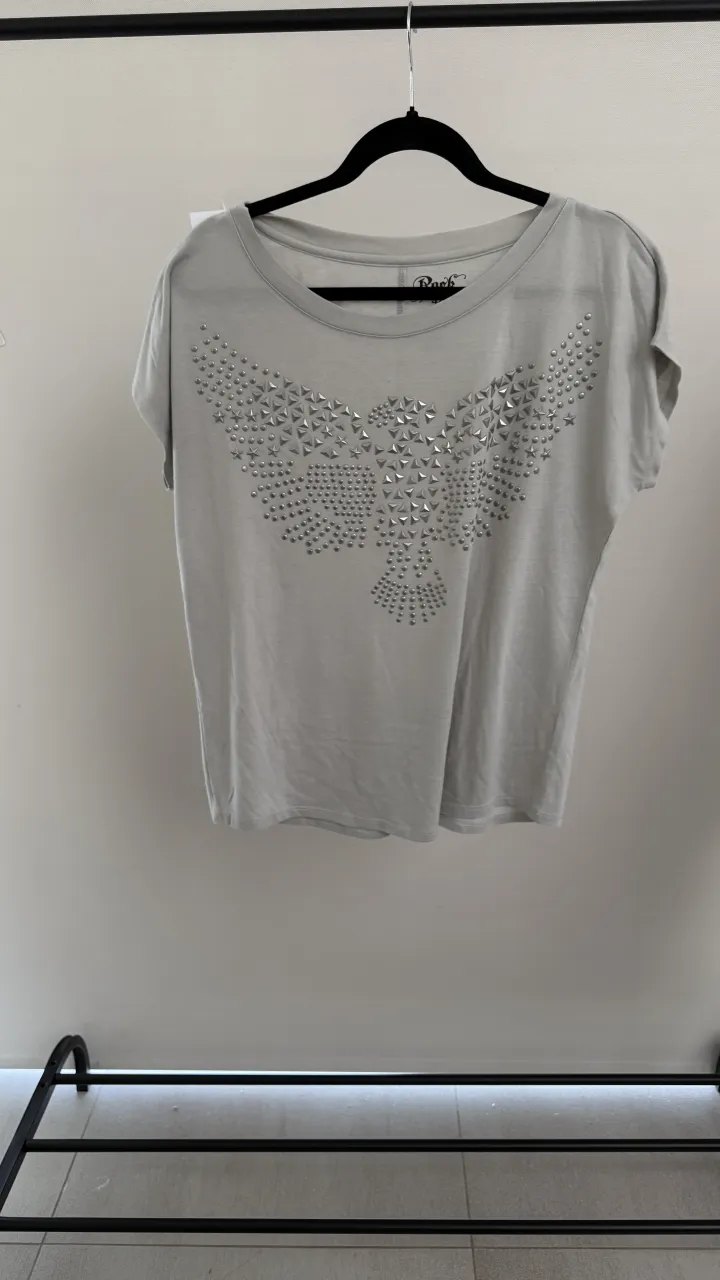 Remera gris con estampa de águila realizada con tachas plateadas en el frente. Cuello redondo y mangas cortas. medidas en fotos