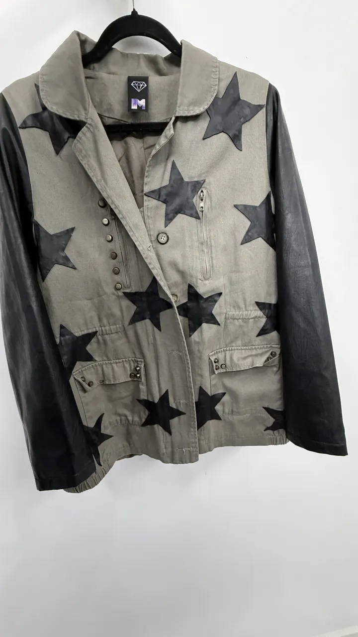 Campera con mangas de cuero sintético y apliques de estrellas negras sobre fondo verde militar. Posee cuello solapa, bolsillos frontales con tapa y cierre, y detalles de botones y tachas. va para un Xs