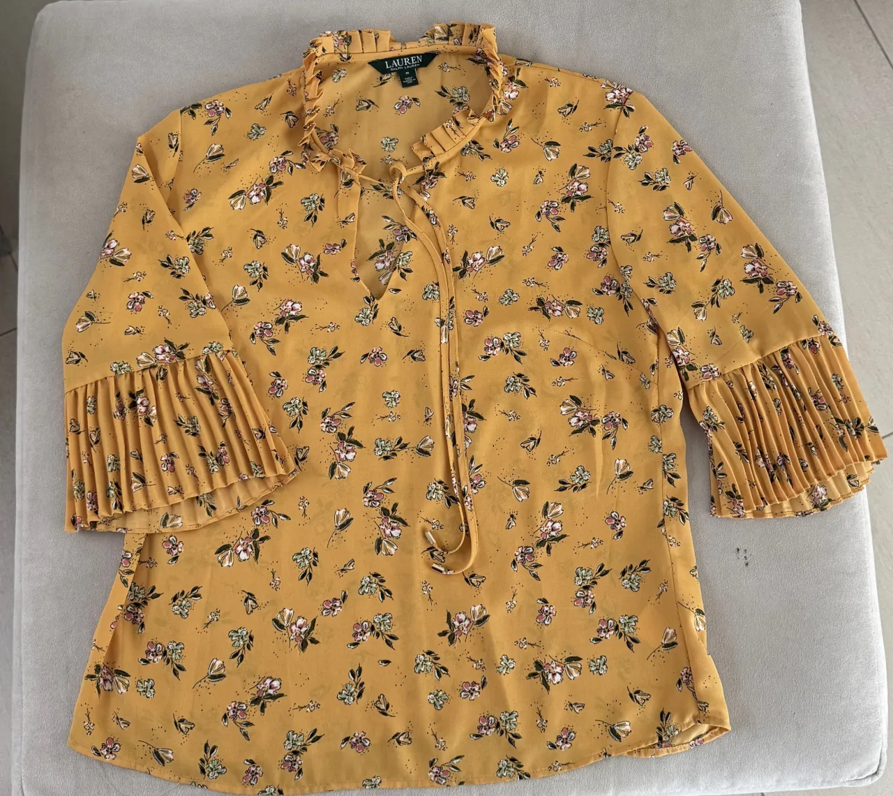 Blusa amarilla con estampa floral de flores pequeñas en tonos rosa, blanco, verde y detalles en negro. Posee cuello mao con volados y lazo para atar. Las mangas son 3/4 con puños plisados y volados.
Marca RALPH LAUREN, talle M. medidas: hombros 38, sisa 49, largo 60
