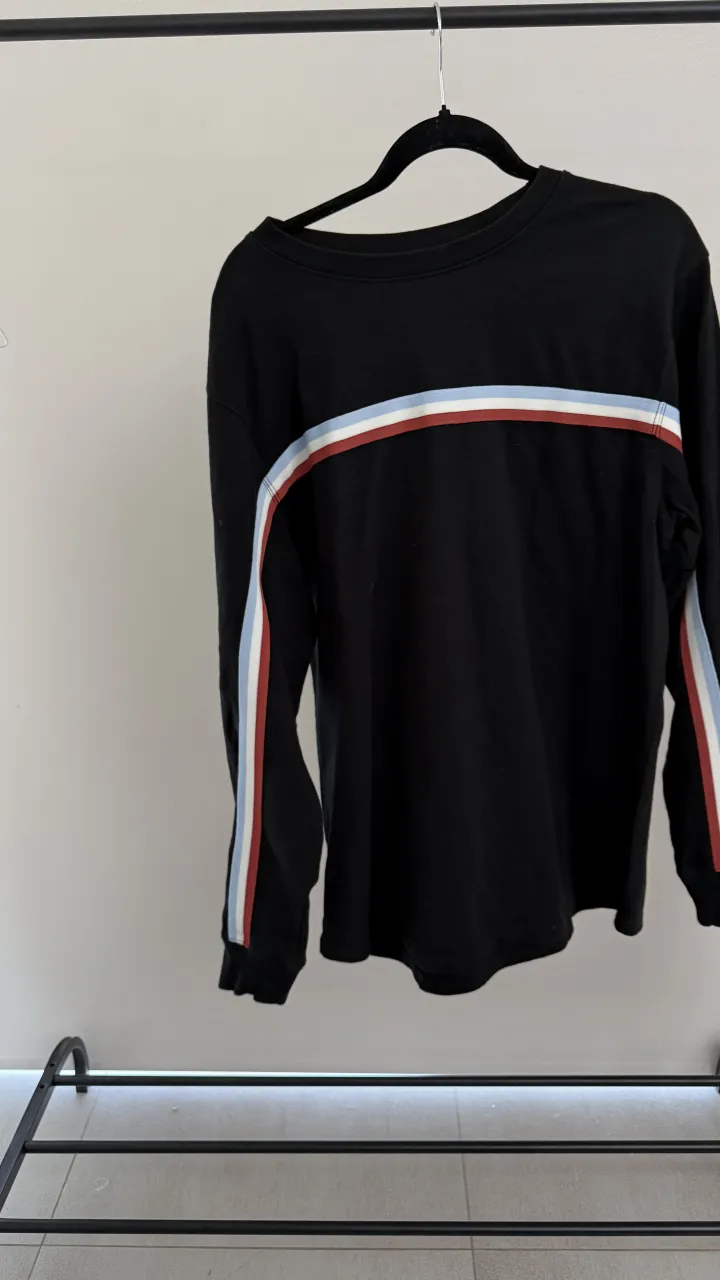 Remera negra de manga larga con detalles de franjas horizontales y verticales en blanco, rojo y celeste. Cuello redondo. medidas en foto