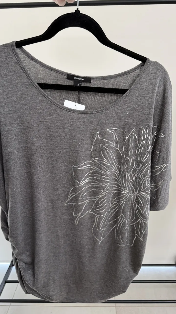 Remera gris con cuello redondo y mangas cortas. Posee un aplique de strass plateado en forma de flor en el frente. Tiene un detalle fruncido con strass en uno de los laterales. medida en fotos