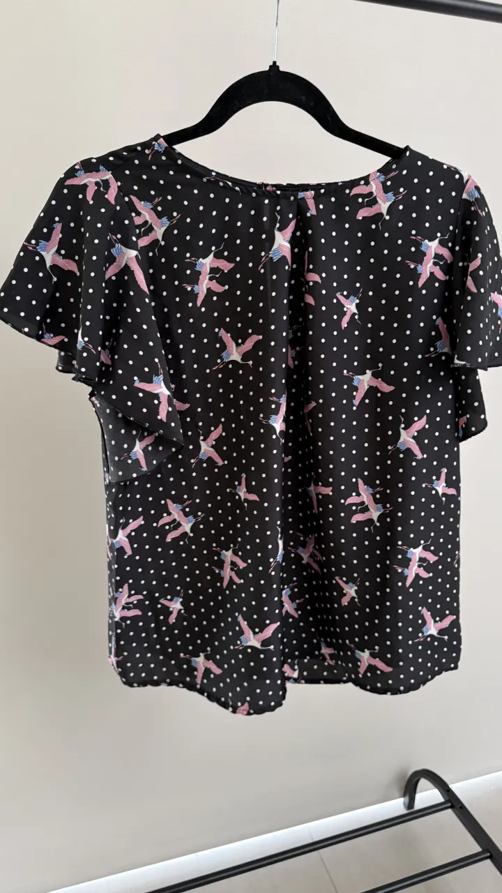 Remera negra con estampa de lunares blancos y flamencos en rosa y celeste. Tiene mangas cortas con volados.