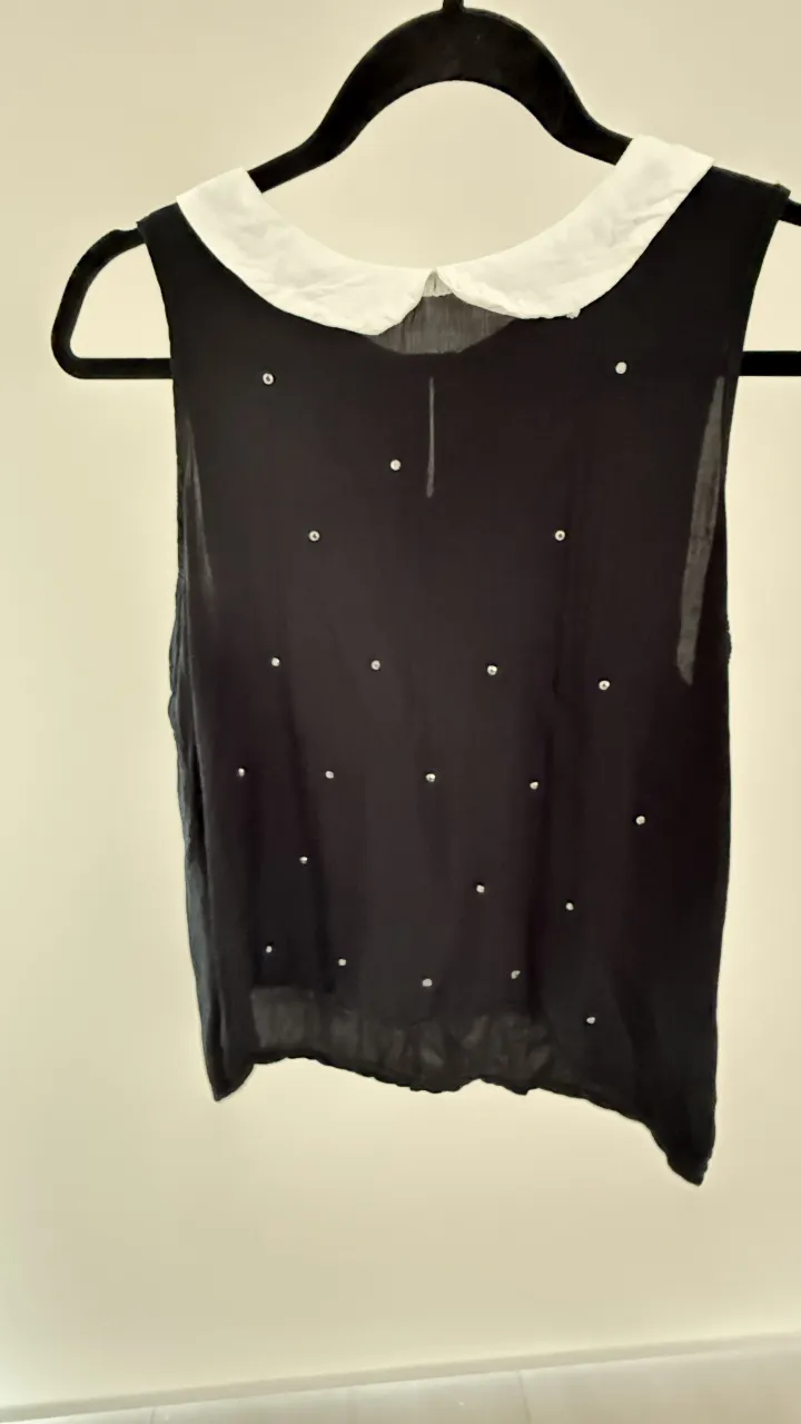 Musculosa negra con cuello blanco y detalles de pequeños apliques plateados distribuidos en el frente. Posee cuello redondo con solapa blanca. 
medidas. hombros 32, sisa 42, largo 55