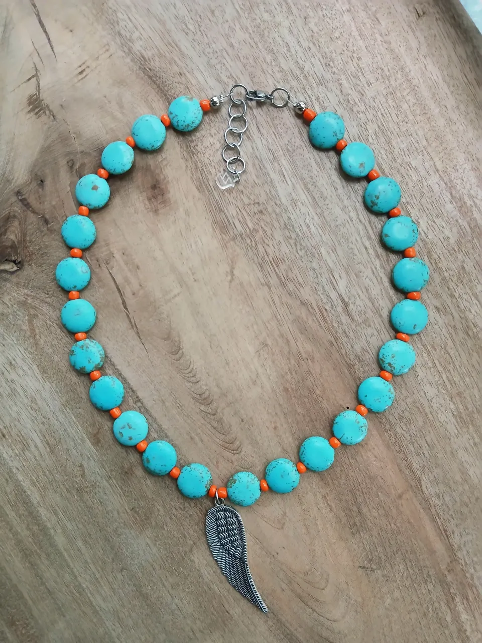 Collar artesanal con piedras turquesas y detalles en naranja. Posee un dije de ala en acero quirúrgico. 