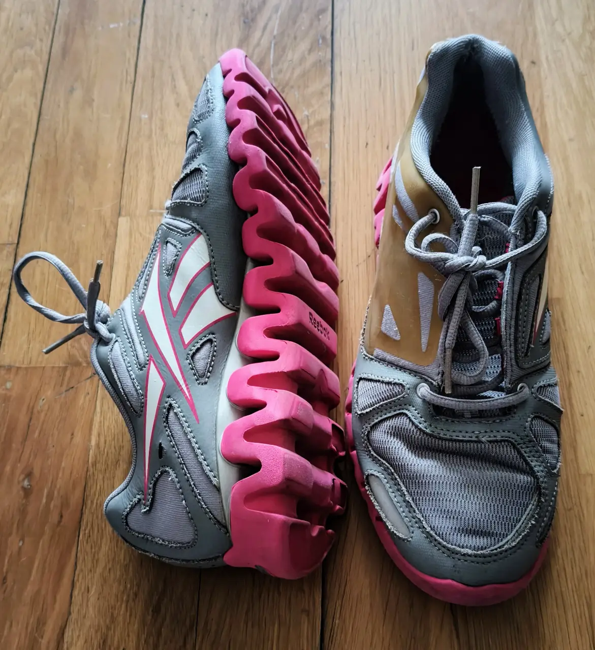 Zapatillas deportivas Reebok para mujer, color gris con detalles en rosa y blanco. Suela con diseño ondulado para mayor amortiguación y estilo.