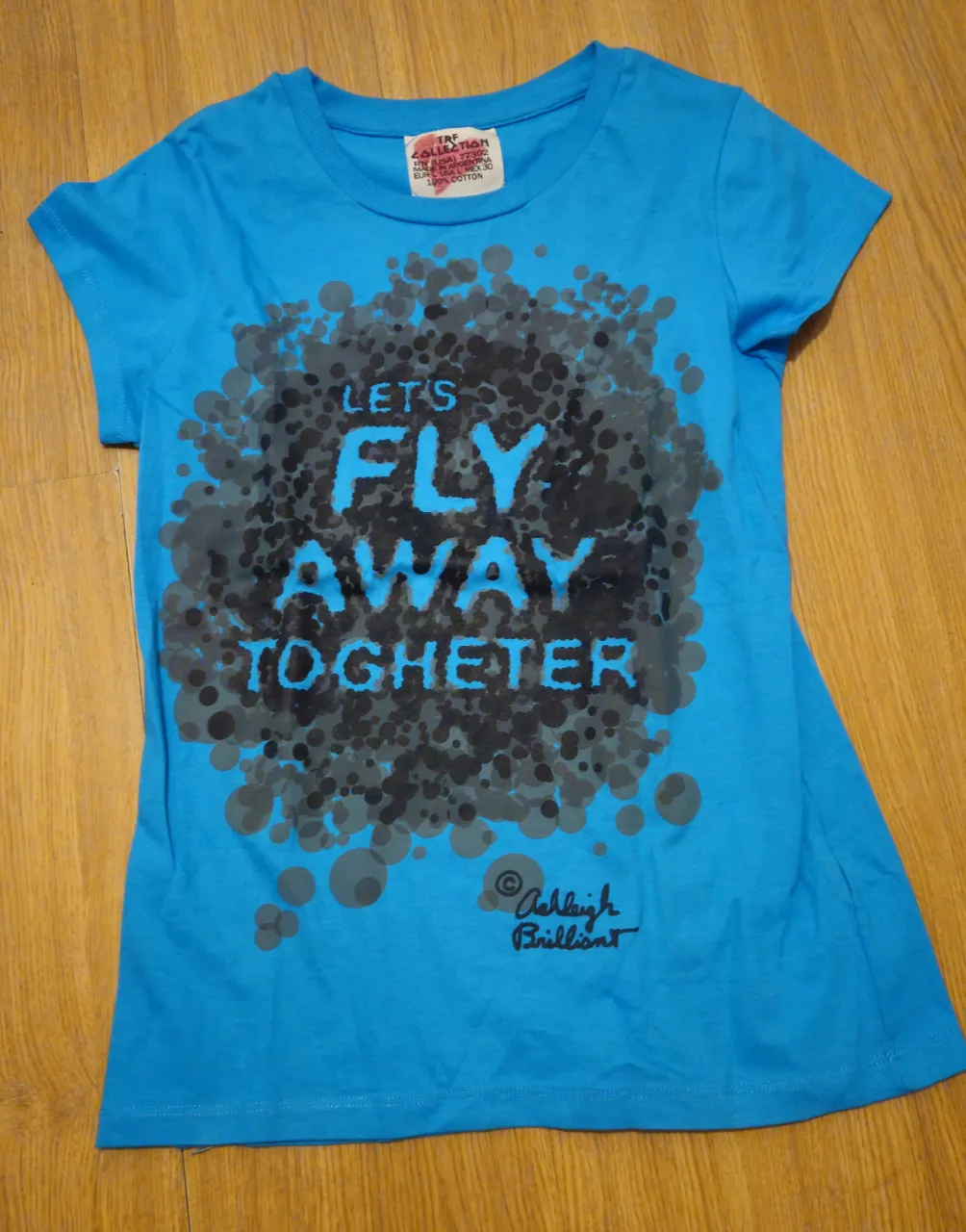 Remera azul de manga corta para niña, con estampado central que dice "LET'S FLY AWAY TOGHETER" en color celeste y gris. Marca Ashleigh Brilliant.