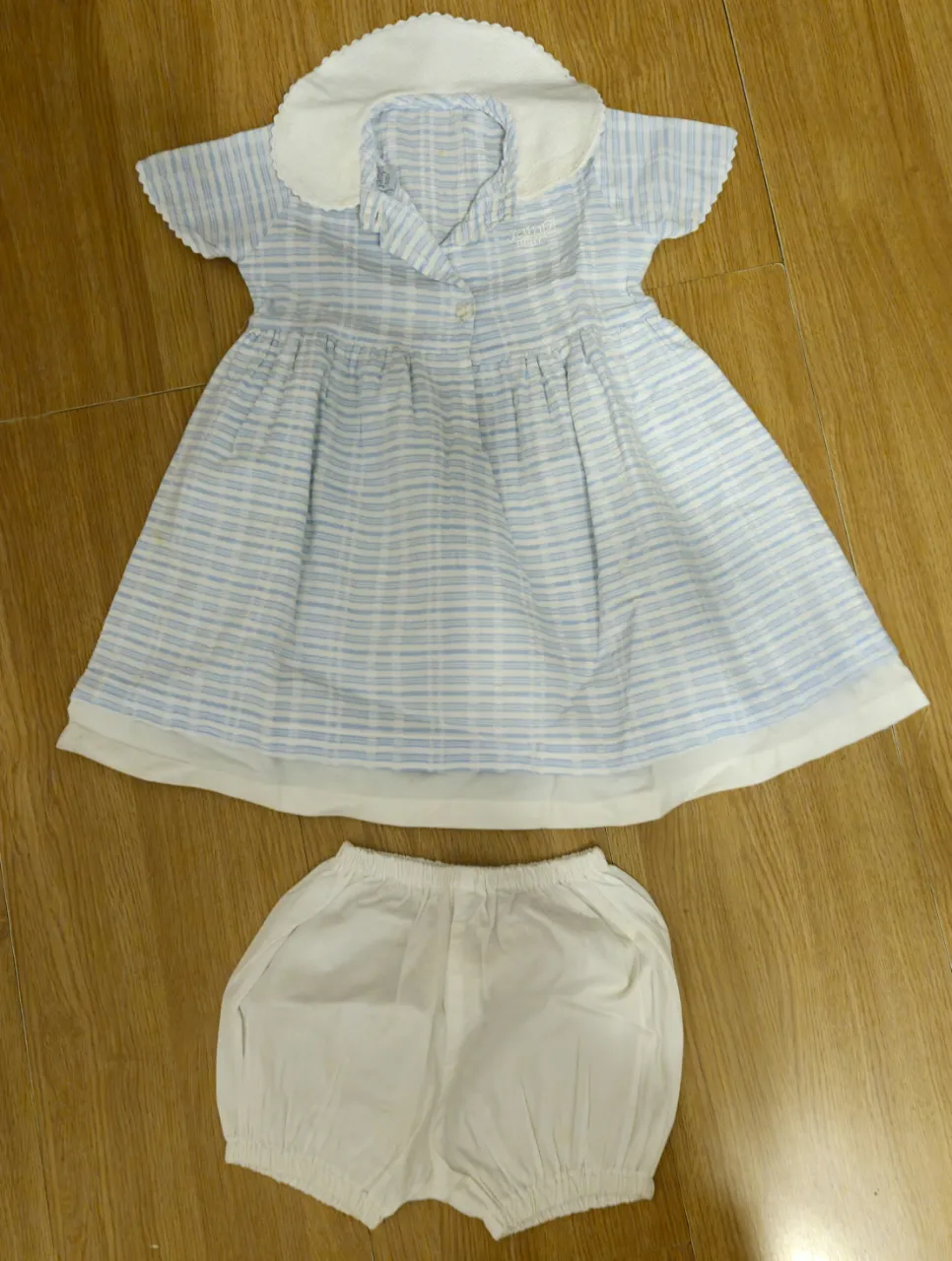 Conjunto de vestido y bombacha para niña, con estampado a rayas celeste y blanco. El vestido tiene cuello bebé y mangas cortas. La bombacha es blanca.