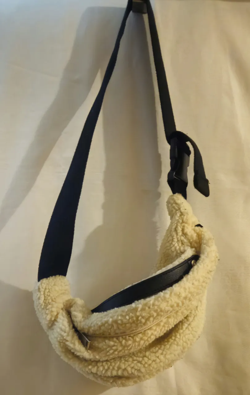 Bandolera tipo riñonera de peluche color crudo con correa negra ajustable. Ideal para llevar lo esencial con estilo y comodidad. Medida 40*22 cm.