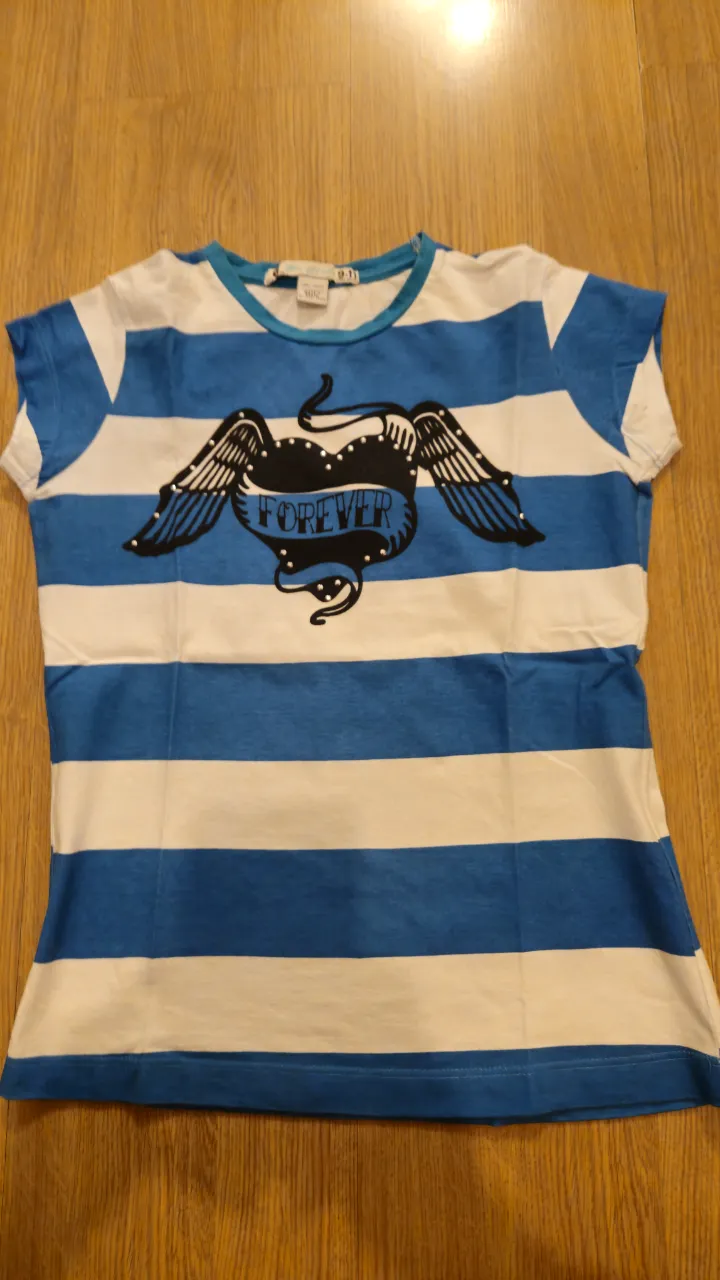 Remera de niña a rayas horizontales blancas y azules, con un diseño de corazón con alas y la palabra "FOREVER" estampada. Cuello redondo con borde azul.