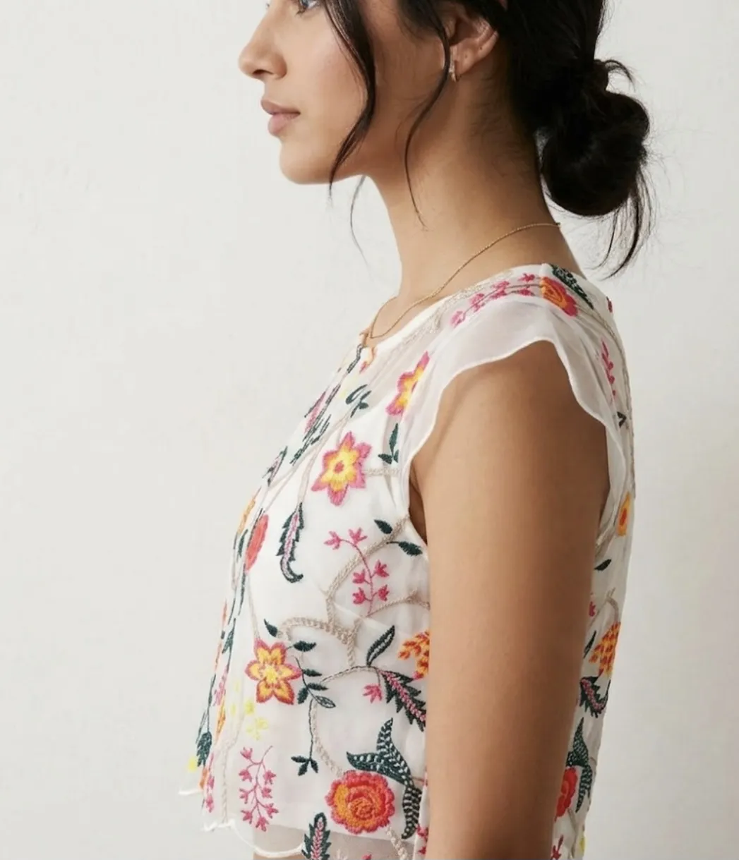 Top blanco con bordado de flores en tonos rosa, naranja, amarillo y verde. Posee mangas cortas y escote redondo.
