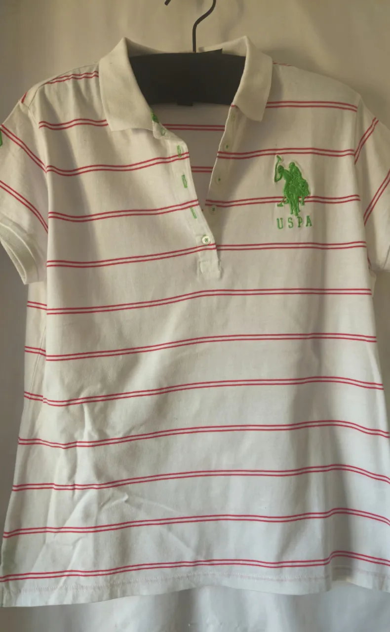 Remera polo de mujer, blanca con rayas rojas horizontales. Tiene el logo de USPA en verde en el pecho y el número 3 en la manga izquierda. Cuello y detalles en verde. Talle XL.