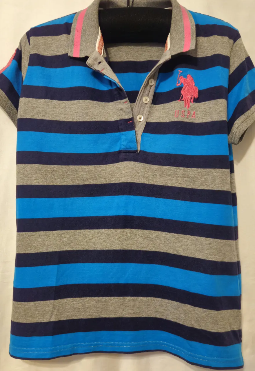 Remera polo de mujer , a rayas horizontales en azul y gris. Con el logo de la marca US Polo Assn. en rosa en el pecho. Cuello y detalles en rosa y gris. Excelente estado.