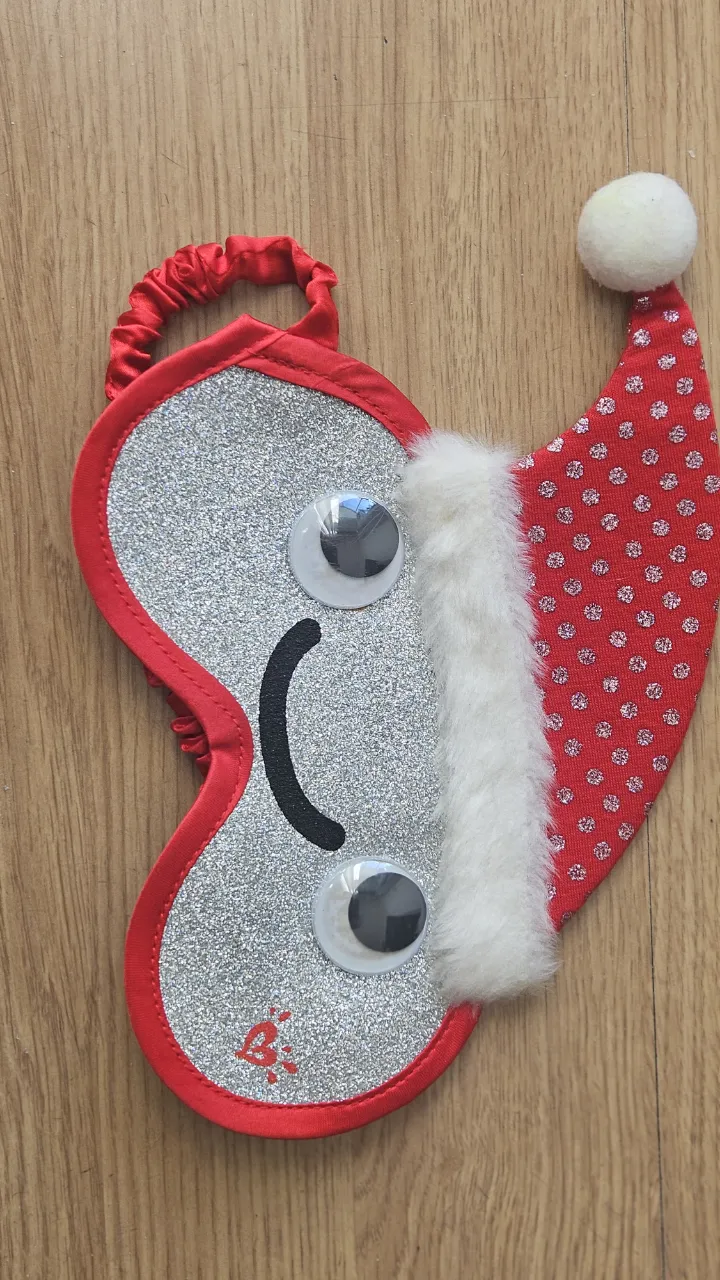 Antifaz navideño con forma de cara, con detalles de gorro de Papá Noel. Ideal para dormir o para usar en fiestas temáticas.