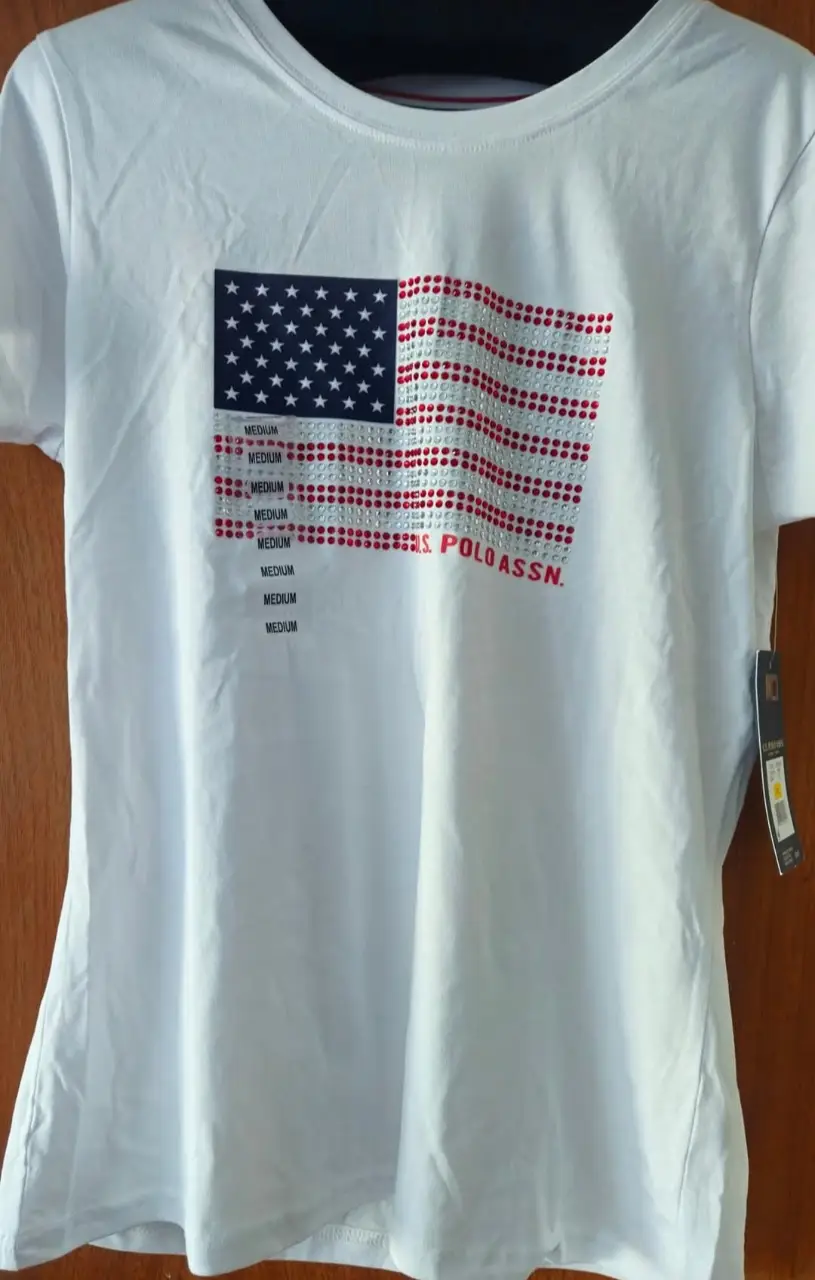 Remera blanca de mujer con estampado de la bandera de Estados Unidos con detalles en strass y el logo de U.S. POLO ASSN. en rojo. Ideal para un look casual y patriótico.