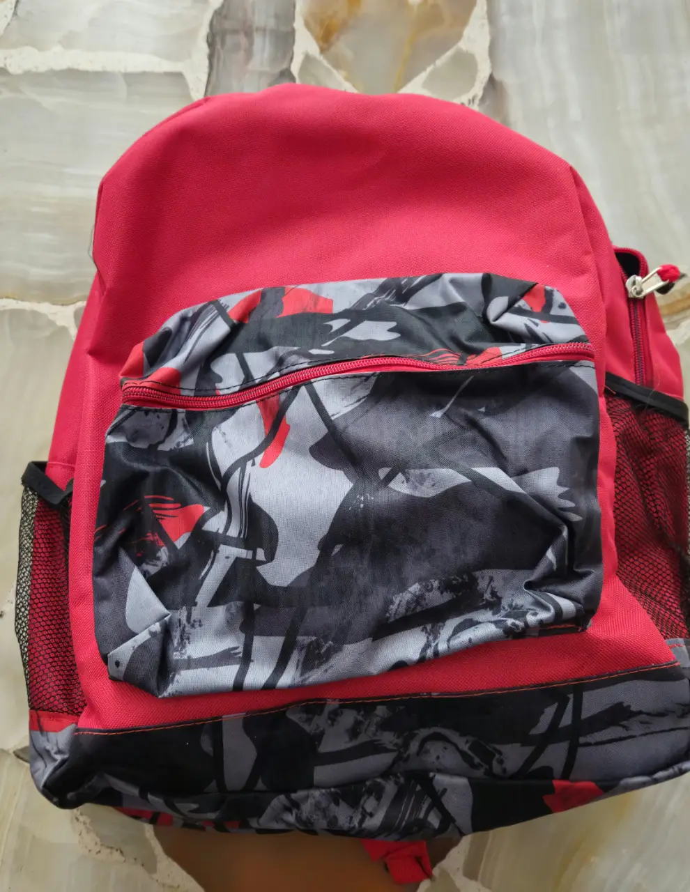 Mochila roja con bolsillo frontal estampado en tonos grises y negros. Ideal para llevar tus cosas con estilo y comodidad. Medida 45*42 cm.