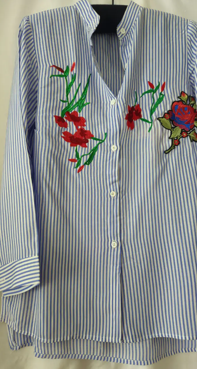 Camisa a rayas celeste y blanca con bordados florales en rojo y verde. Cuello mao y botones frontales. Ideal para un look casual y fresco.