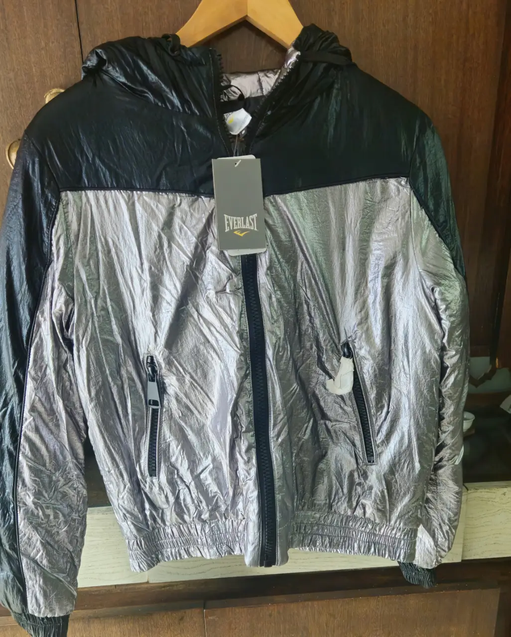 Campera Everlast deportiva con capucha, combinada en negro y plateado. Con cierre frontal y bolsillos con cierre. Ideal para actividades deportivas o uso casual.