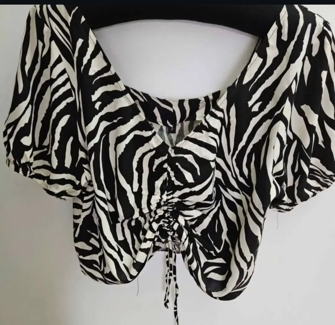 Blusa crop con estampado de cebra en beige y negro. Diseño con escote en V y detalle fruncido en el centro. Mangas cortas abullonadas. Ideal para un look moderno y audaz.