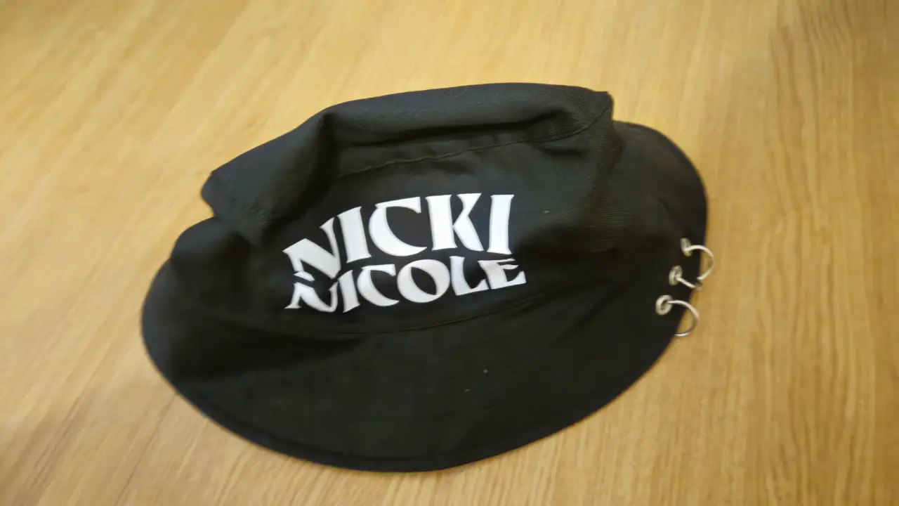 Gorra bucket negra con el nombre "NICKI NICOLE" en blanco. Tiene detalles de aros plateados en el borde. Disponible 5 unidades.