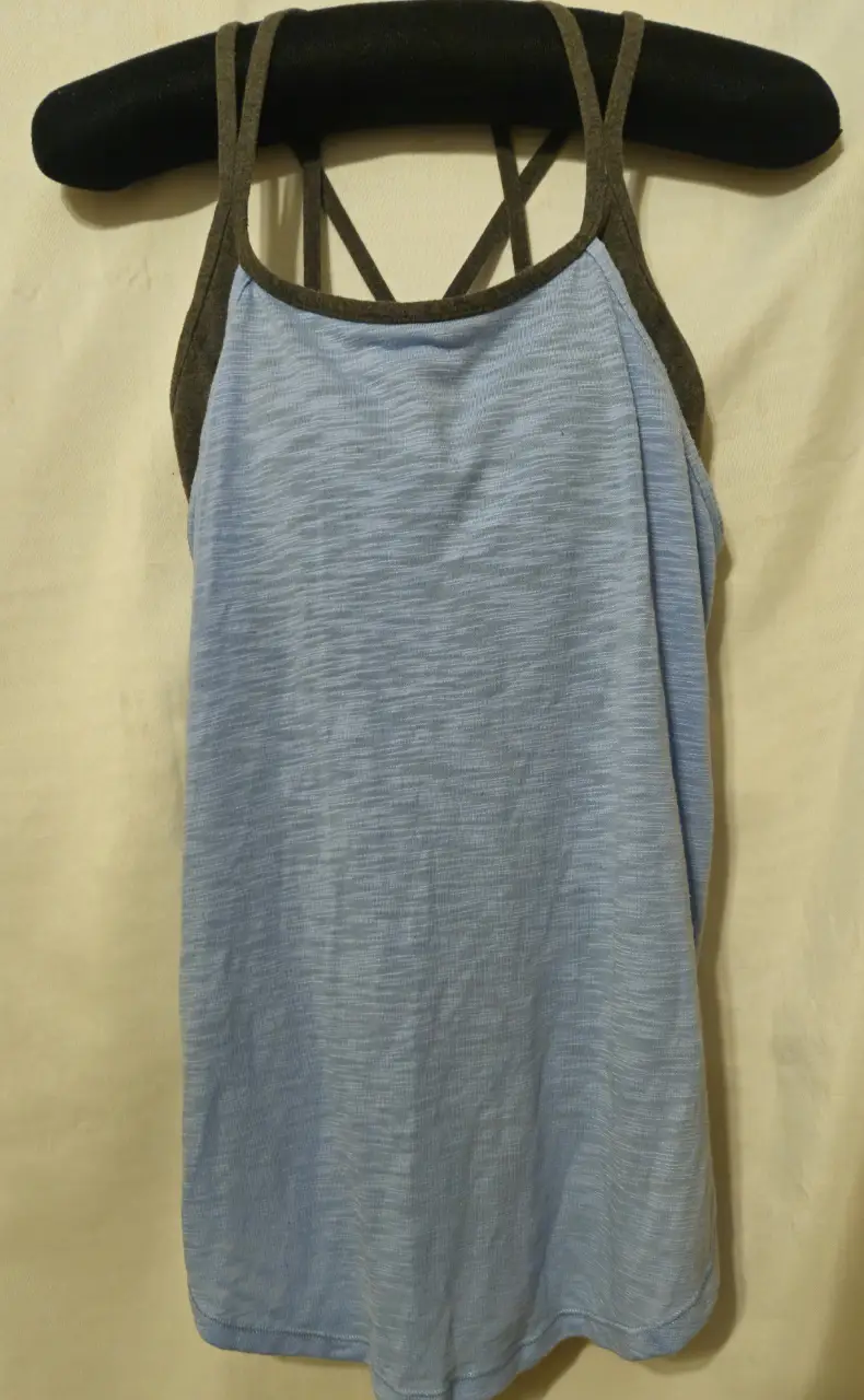 Musculosa deportiva de mujer, con diseño de espalda cruzada y doble capa. La capa interior es de color azul claro, mientras que la exterior es gris oscuro. Ideal para actividades físicas.