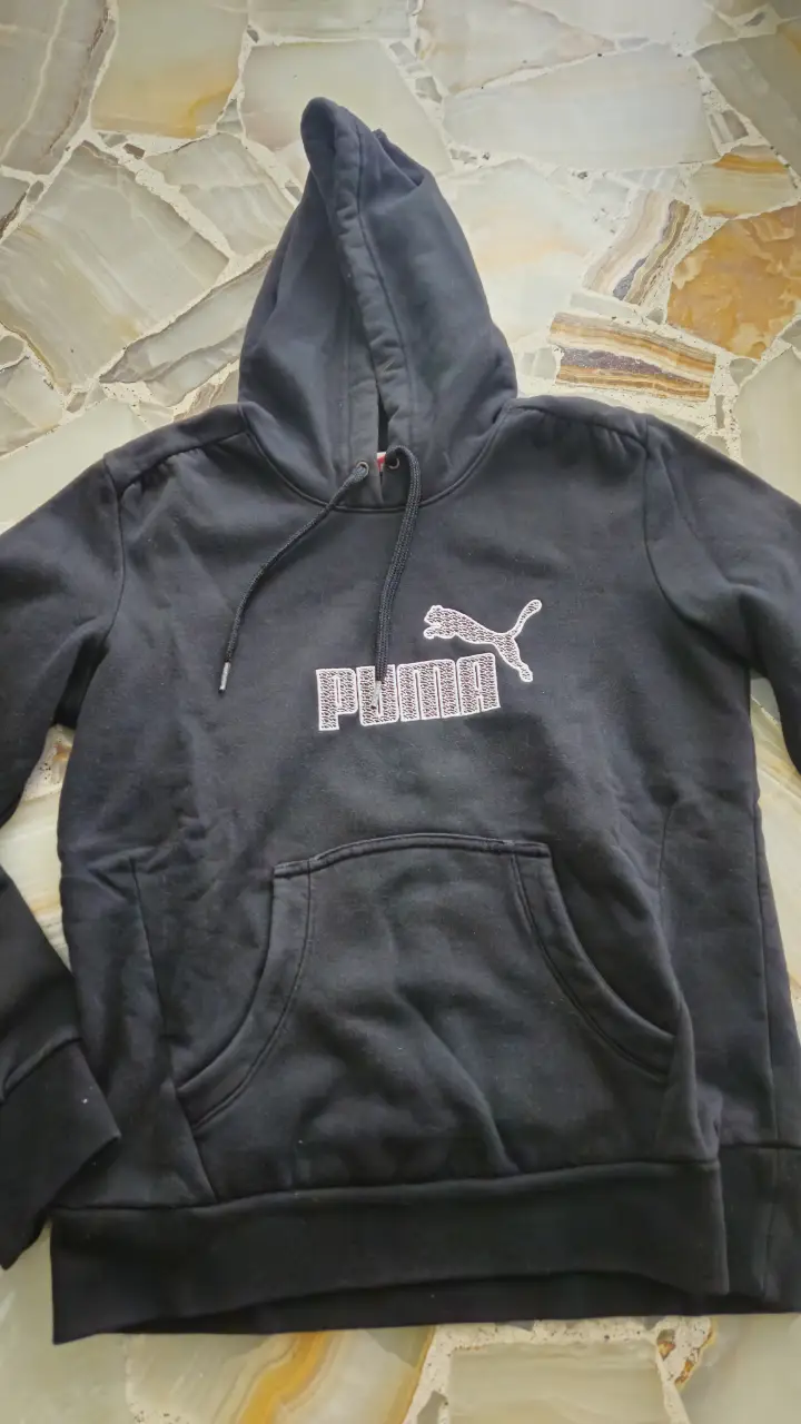 Buzo canguro negro de hombre, con capucha y bolsillo frontal. Presenta el logo de Puma en blanco en el pecho. Ideal para un look casual y cómodo.