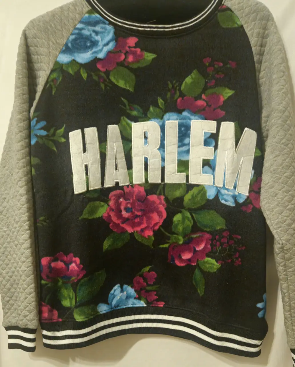 Buzo de cuello redondo con estampado "HARLEM" en letras blancas y flores en tonos rosa y azul. Mangas grises con diseño acolchado. Detalles de rayas blancas y negras en el borde inferior y puños.