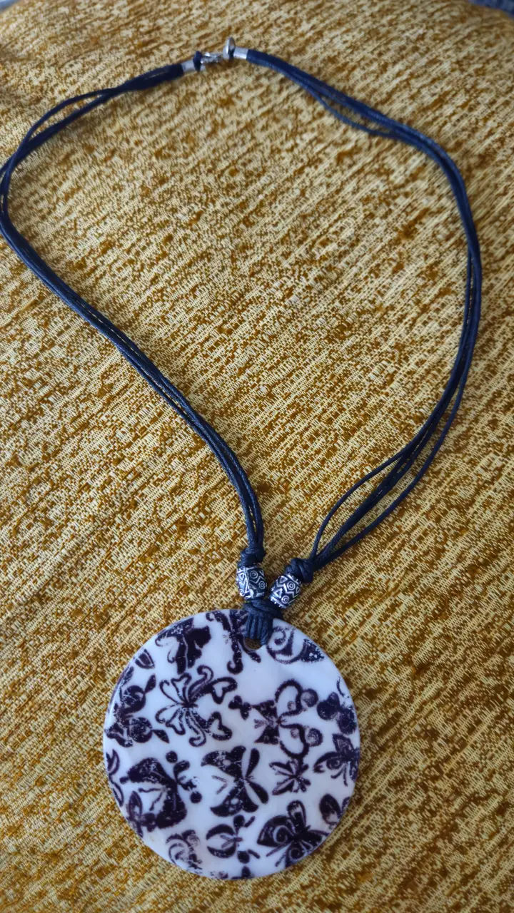 Collar artesanal con dije redondo de cerámica con diseño floral en blanco y negro. El collar tiene un cordón negro.
