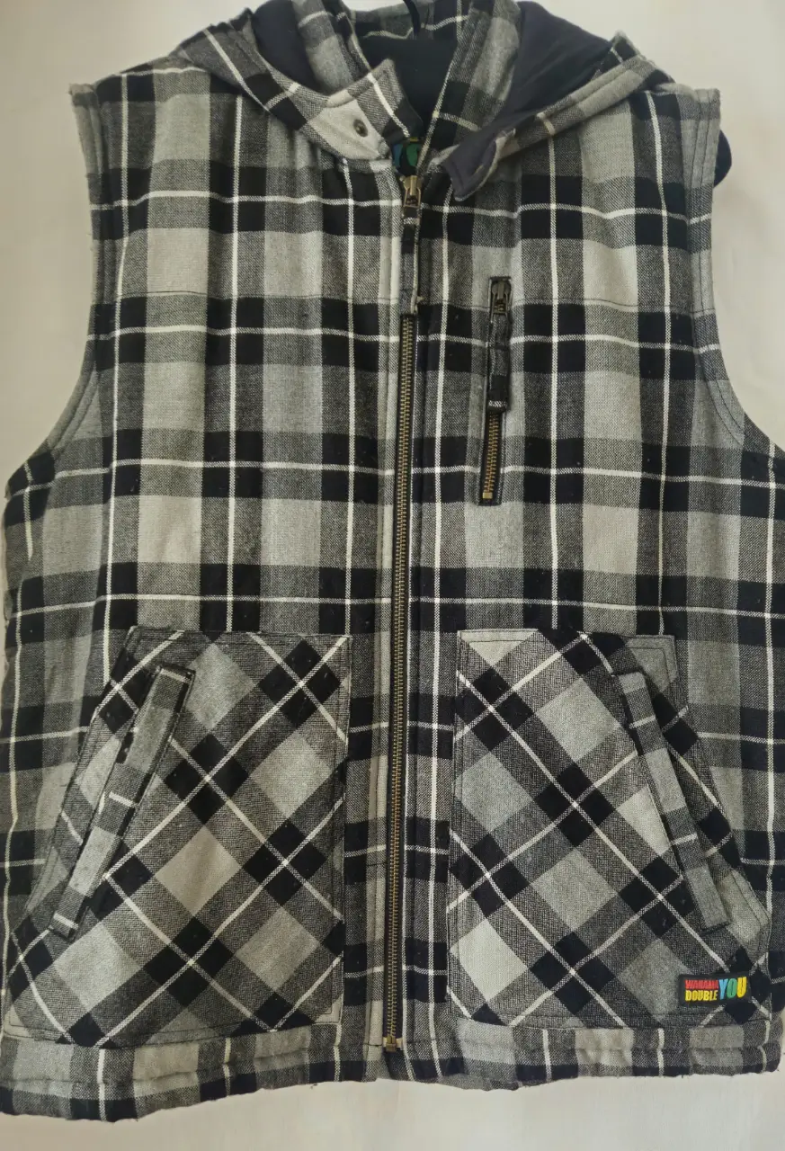 Chaleco con capucha para hombre, de tela a cuadros en tonos grises y negros. Tiene cierre frontal, bolsillos delanteros y un pequeño bolsillo con cierre en el pecho. Ideal para un look casual y abrigado.