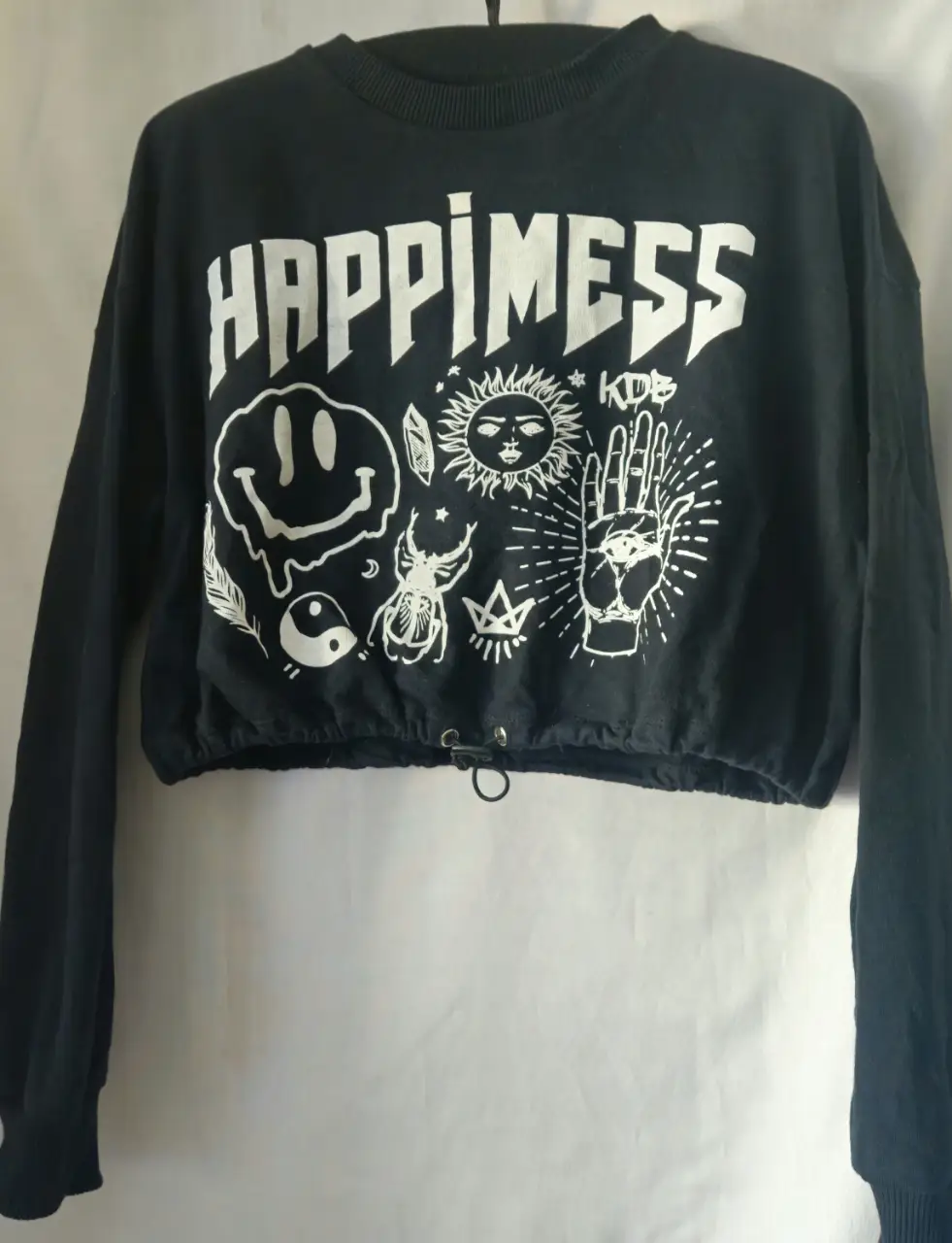 Buzo crop negro con estampado "Happimess" en blanco. Diseño con dibujos y letras, ideal para un look urbano y moderno. Cuenta con cordón ajustable en la cintura y mangas largas.