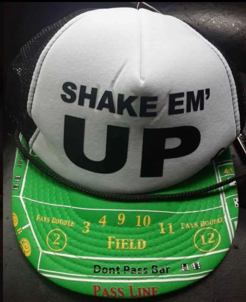 Gorra trucker con diseño de mesa de craps. La parte frontal es blanca con la frase 'Shake Em' Up' en negro. La visera es verde con el diseño de la mesa de craps. La parte trasera es de malla negra. Ideal para un look casual y divertido.