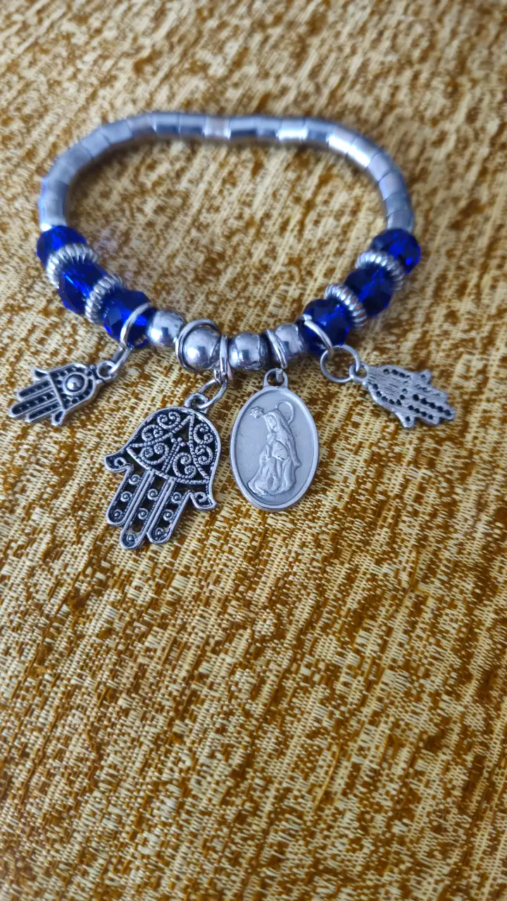 Pulsera artesanal con cuentas azules y dijes plateados, incluyendo manos de Fátima y un dije de la Virgen.
