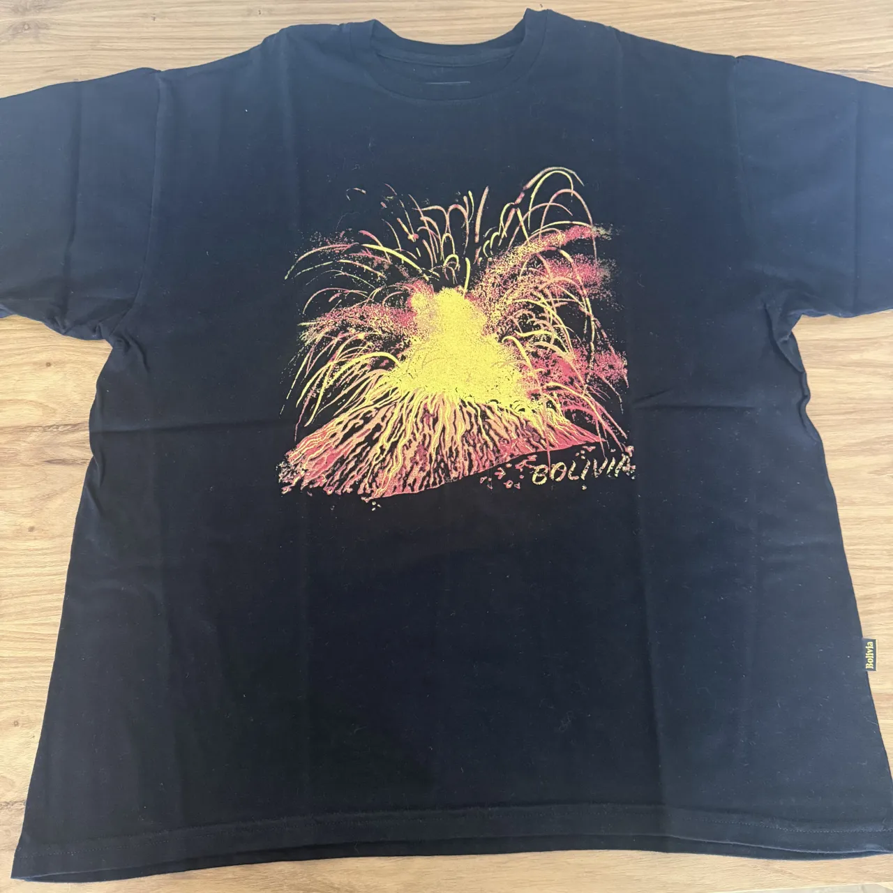 Remera DIVINA BOLIVIA negra con estampa de un volcán en erupción con fuegos artificiales en colores amarillo, rosa y rojo. La palabra "BOLIVIA" se lee debajo de la estampa.