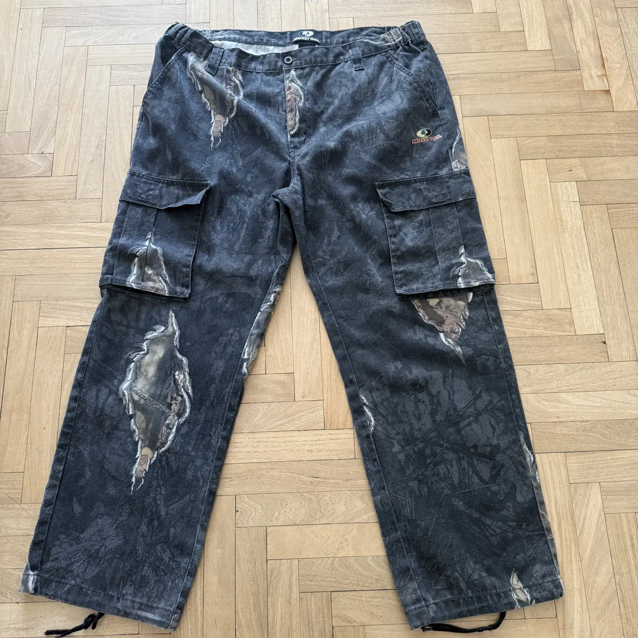 Pantalones MOSSY OAK cargo con estampado camuflado y roturas. Tienen bolsillos cargo en los laterales y cordones en los tobillos.