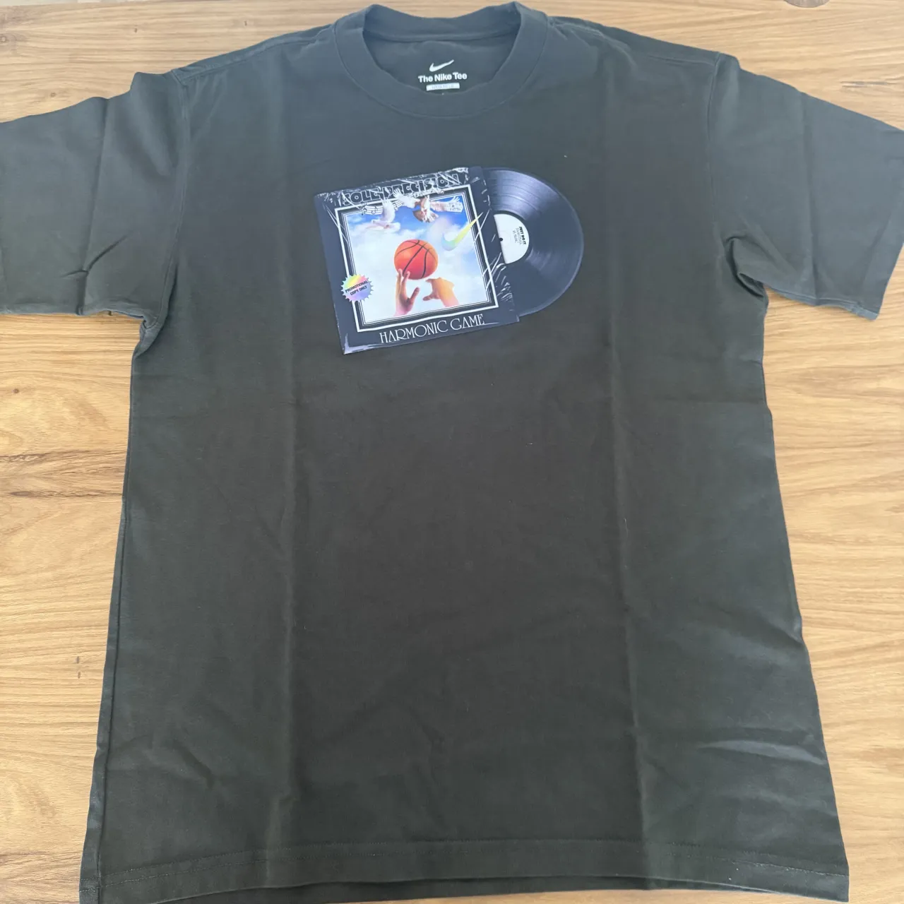 Remera NIKE de manga corta de color verde oscuro con estampa de un disco de vinilo y una imagen alusiva a "Space Jam" con el texto "HARMONIC GAME". La marca Nike es visible en la etiqueta del cuello.