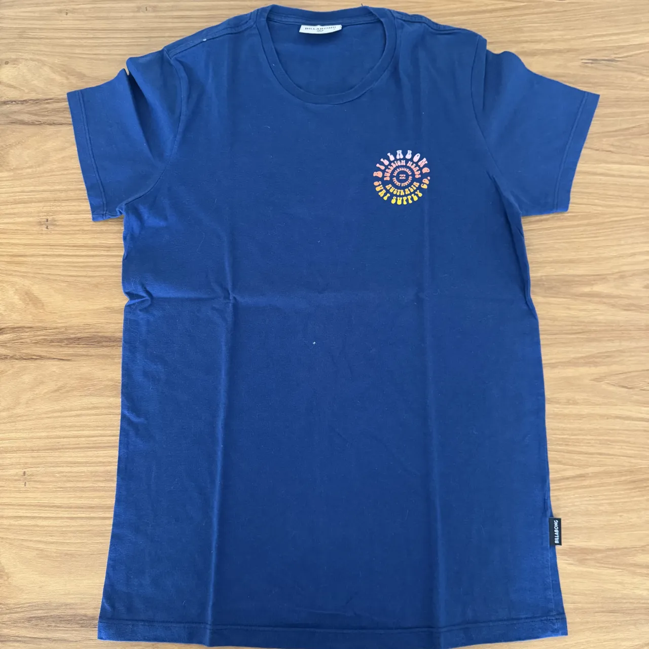 Remera azul de manga corta con estampa circular multicolor en la espalda y logo pequeño en el frente. Marca visible en etiqueta.