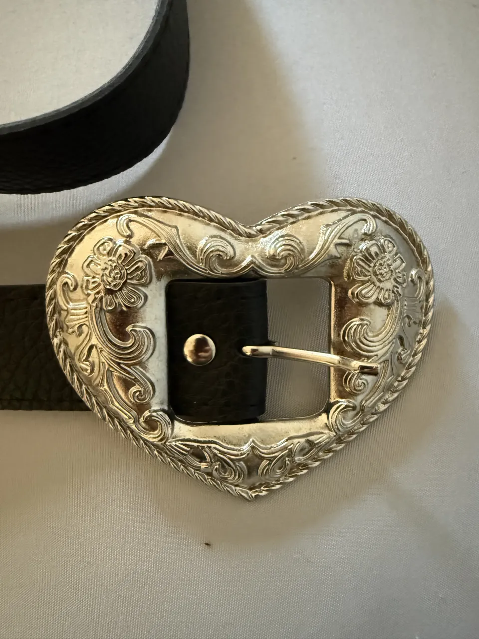 Cinturón negro con hebilla plateada en forma de corazón con detalles florales y borde trenzado.