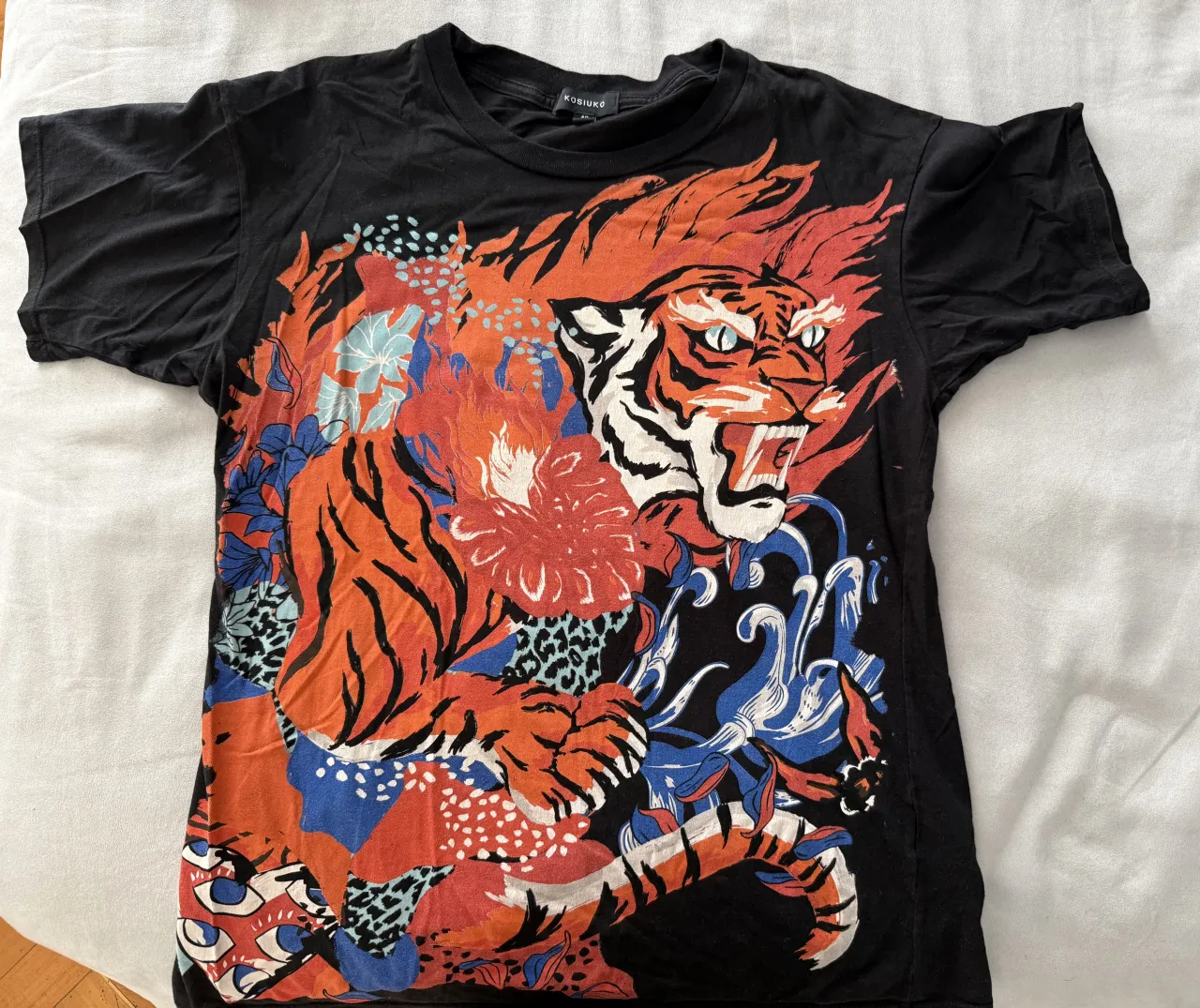 Remera negra con un impactante estampado de tigre rugiendo rodeado de flores y elementos abstractos en tonos vibrantes de naranja, rojo, azul y blanco. Un diseño audaz y llamativo, ideal para quienes buscan destacar con un estilo único.