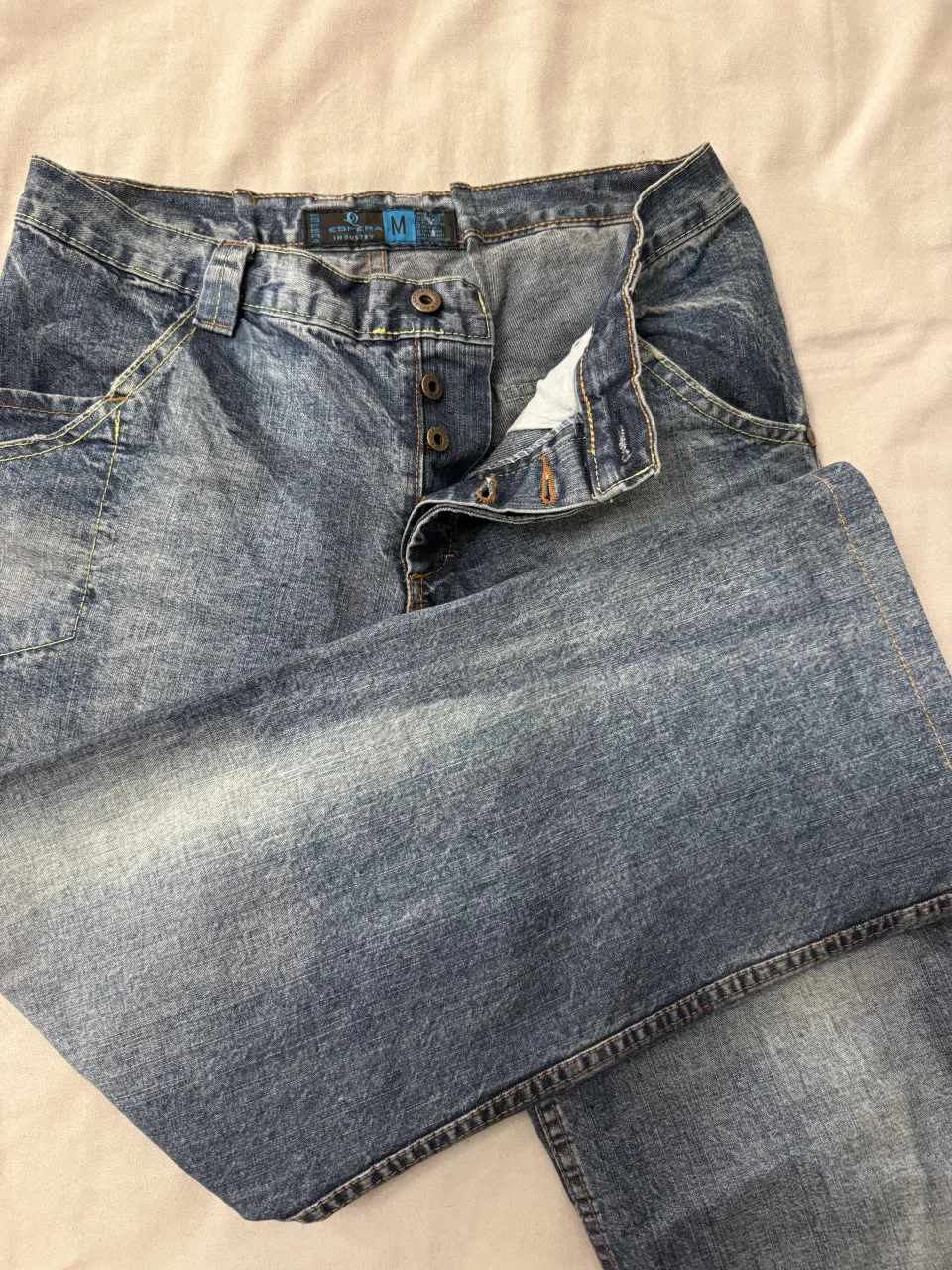 Jeans de corte clásico con detalles de desgaste y botones frontales. Presenta bolsillos delanteros y traseros con detalles de costura y un parche distintivo en la parte trasera.