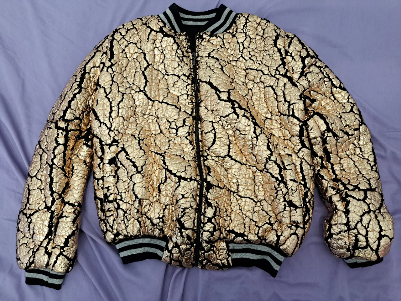 Campera bomber dorada con diseño craquelado. Ideal para un look de noche. Detalles en negro y rib en cuello, puños y cintura. (Largo 55cm, ancho 48cm)