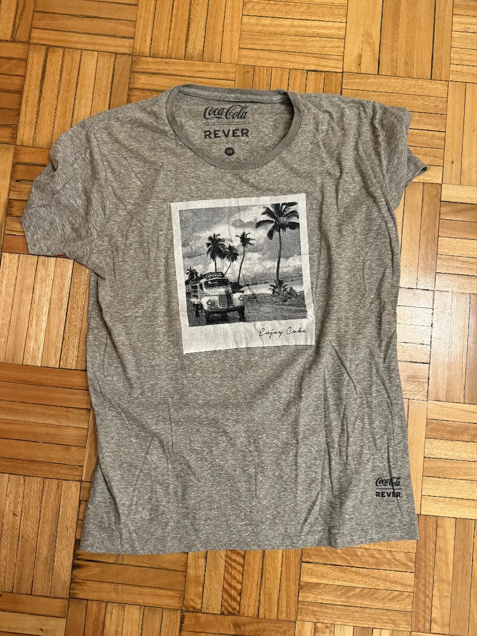 Remera de hombre gris con estampado de Coca-Cola. Diseño retro con imagen de camioneta en la playa y la frase 'Enjoy Coke'. Cuello redondo y manga corta. Ideal para un look casual y con onda.