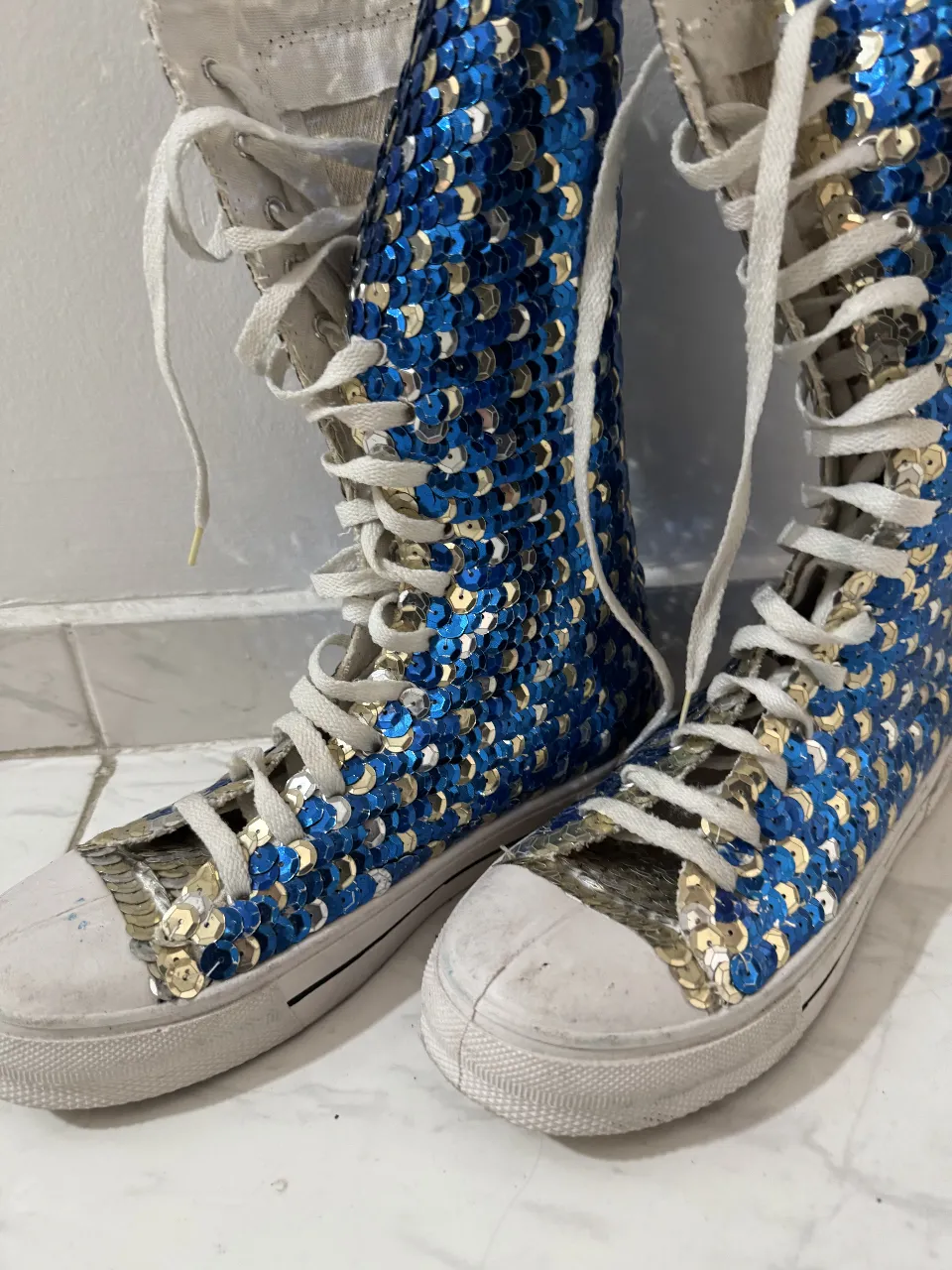 Botas altas de lona con lentejuelas azules y doradas. Ideales para un look de fiesta o para destacar en eventos especiales. Tienen cordones blancos y suela blanca.