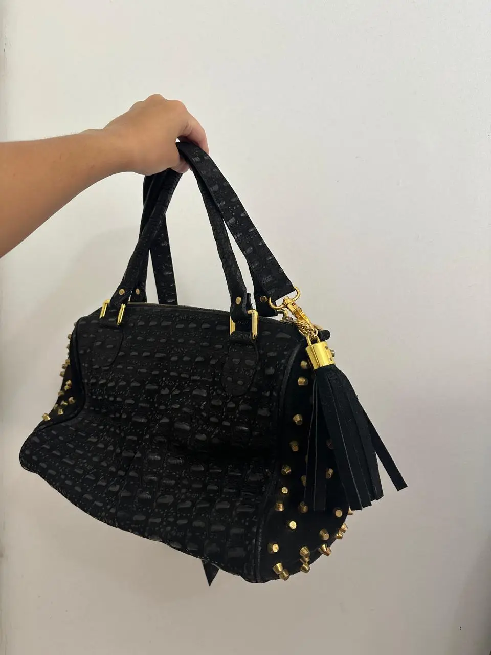 Cartera negra con textura de cocodrilo y tachas doradas. Incluye correa para llevar al hombro y manijas cortas. Ideal para un look elegante y moderno.