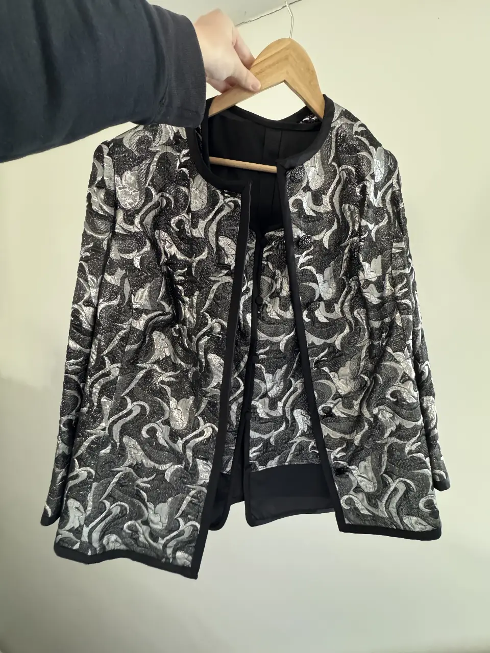 Elegante blazer de mujer con estampado floral en tonos plateados y negros. Diseño abierto con detalles en negro y botones decorativos. Ideal para eventos formales o para darle un toque sofisticado a tu look. Viene con una remera dentro