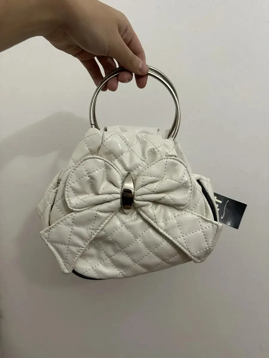 Cartera blanca con forma de moño y detalles acolchados. Tiene manija de metal y correa para colgar.