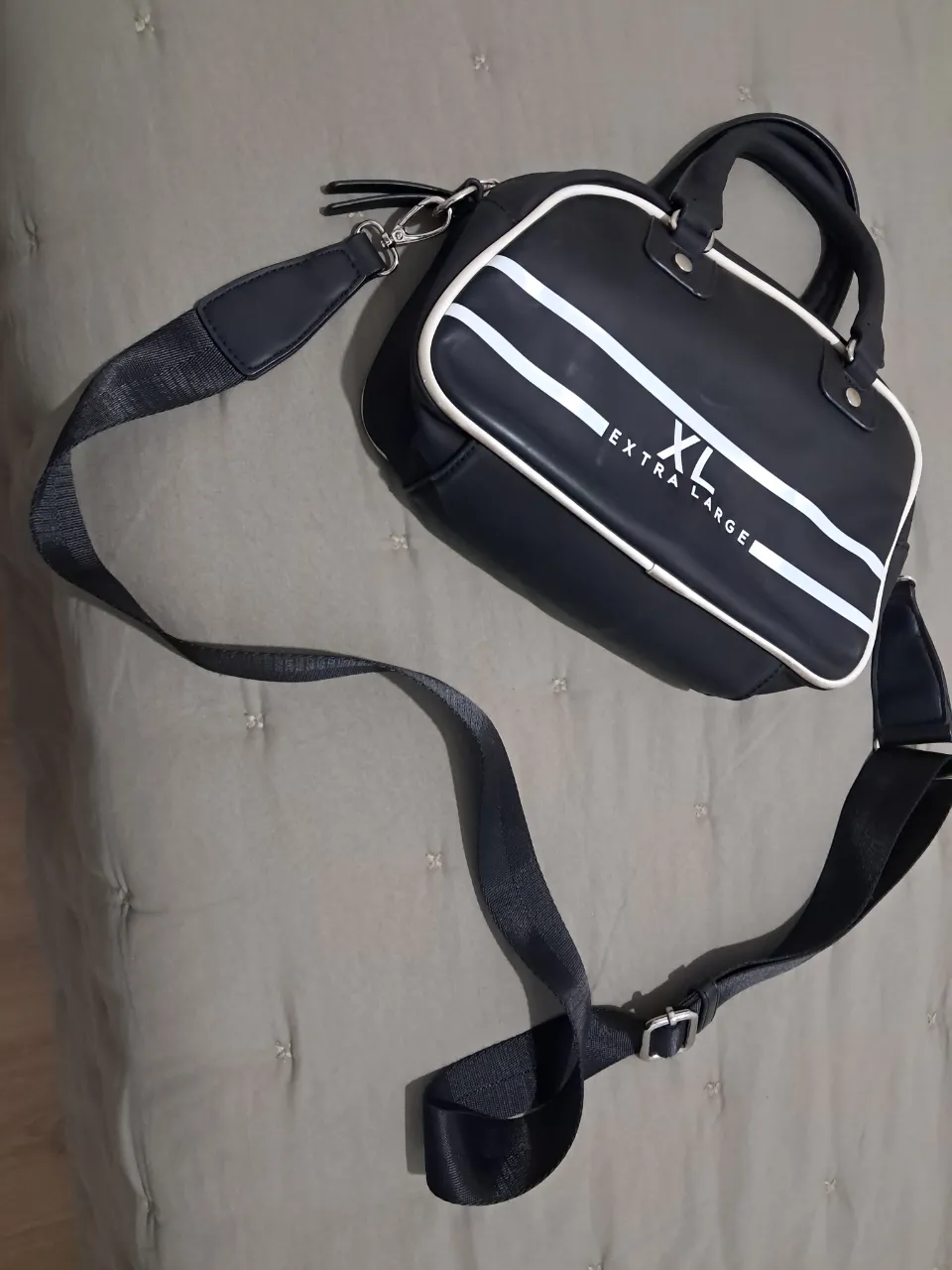 Bolso negro con detalles blancos y logo XL Extra Large. Tiene dos asas cortas y una correa larga desmontable.