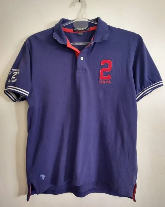 Remera POLO azul con detalles en rojo y blanco. Posee un número 2 bordado en el frente y en la manga izquierda, junto con el logo de la marca. Ideal para un look casual y deportivo.