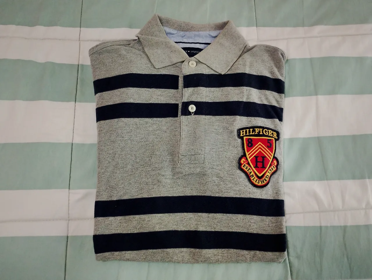 Remera polo de manga corta a rayas grises y azul marino. Tiene un escudo bordado de la marca en el pecho. Ideal para un look casual y moderno.