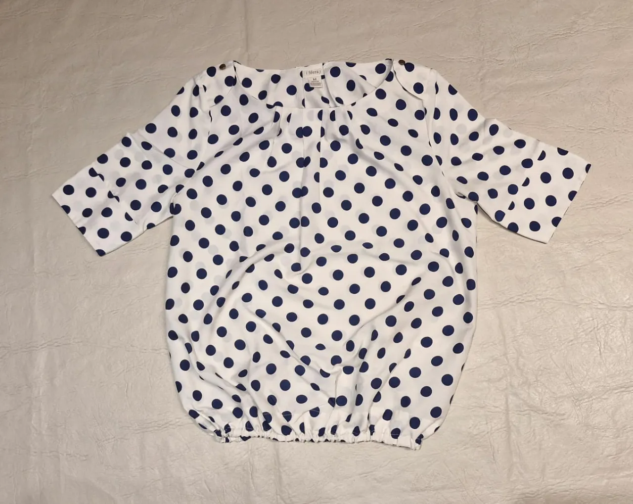 Blusa de mujer con estampado de lunares azules sobre fondo blanco. Cuello redondo, con frunces, mangas 3/4 y elástico en la cintura. Con detalle de sobremesa y botón dorado en los hombros. Ideal para un look casual y femenino.
